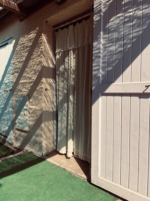 A building with a textured exterior wall, a wooden door with a white curtain, and a green artificial turf area. - Fisherman's Island Mas (Le Barcarès)