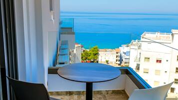 A balcony with a round table and two chairs, overlooking a cityscape and the sea.