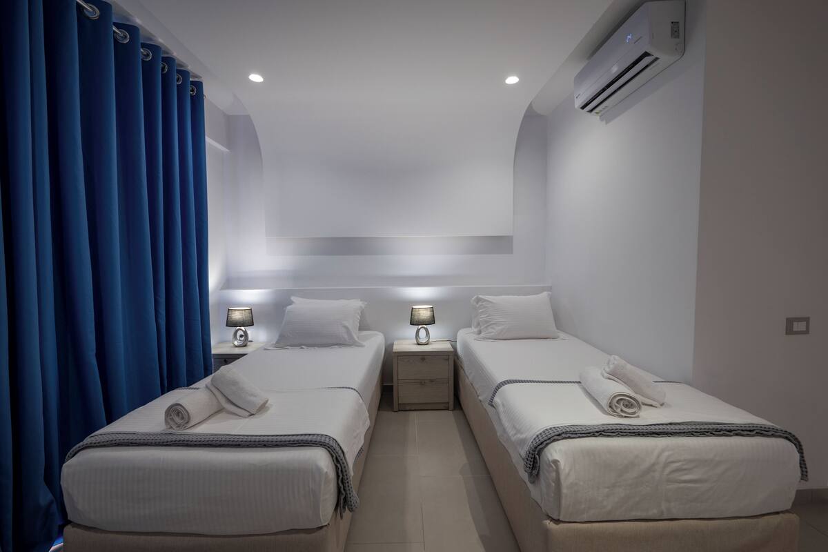 A hotel room with two beds, a nightstand with a lamp, an air conditioner, and a curtain.
