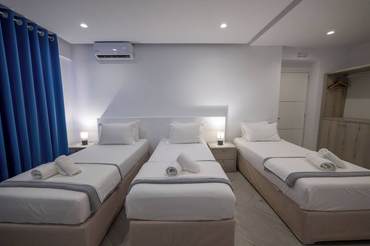 A hotel room with three single beds, a wardrobe, a bedside table with a lamp, and an air conditioning unit.