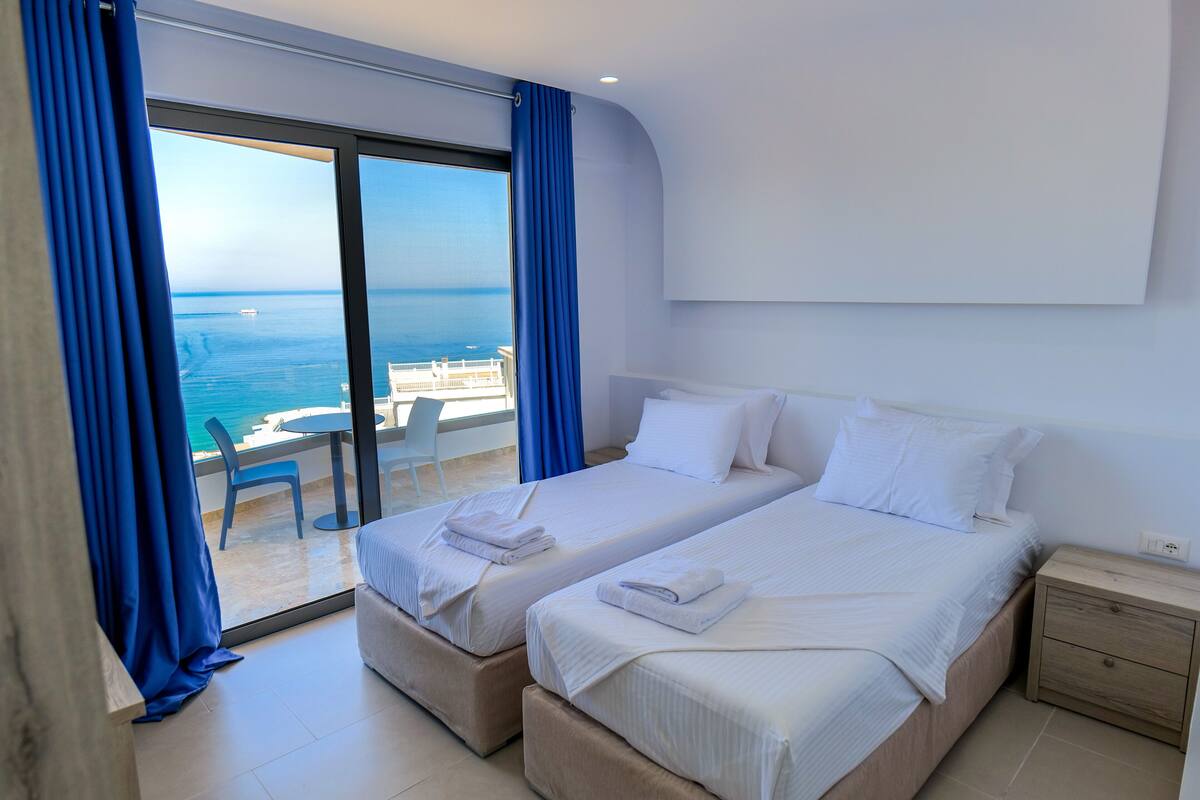 A hotel room with two beds, a balcony with a table and chairs, and a view of the sea.