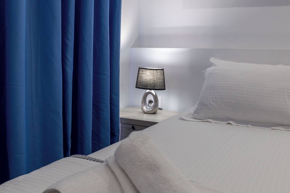 A neatly made bed with white linens, a rolled white towel, a bedside lamp, and blue curtains.