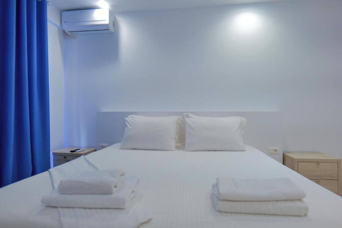 A hotel room with a bed, pillows, a nightstand, an air conditioning unit, and a blue curtain.