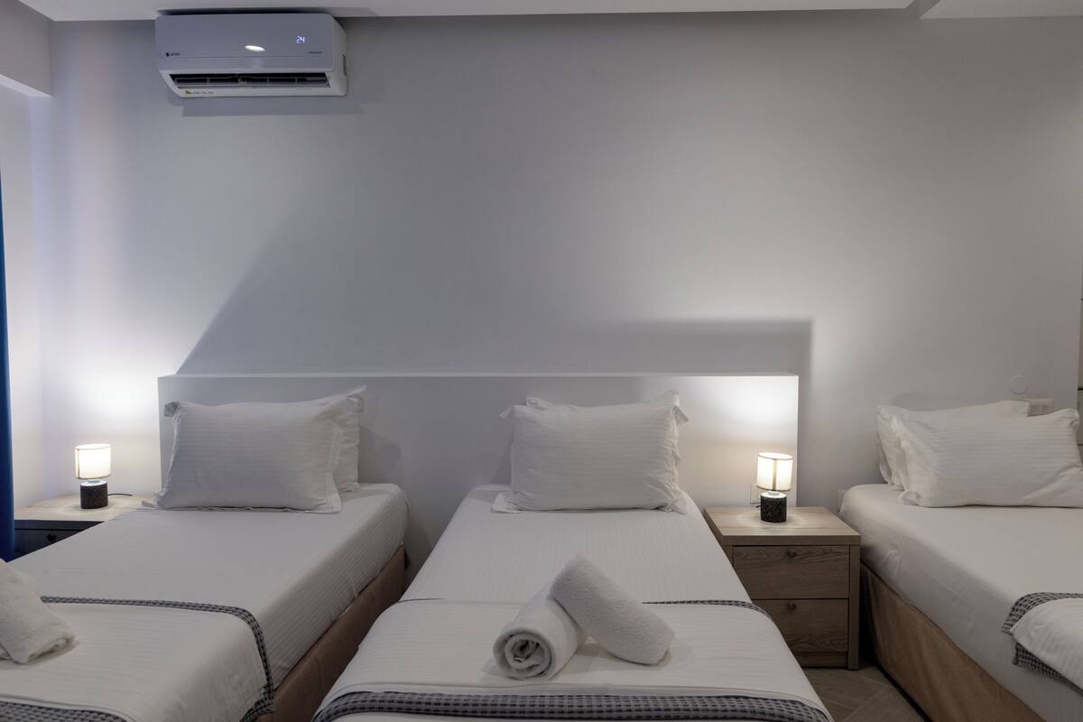A hotel room with two beds, a nightstand with a lamp, a wall-mounted air conditioner, and a ceiling light.