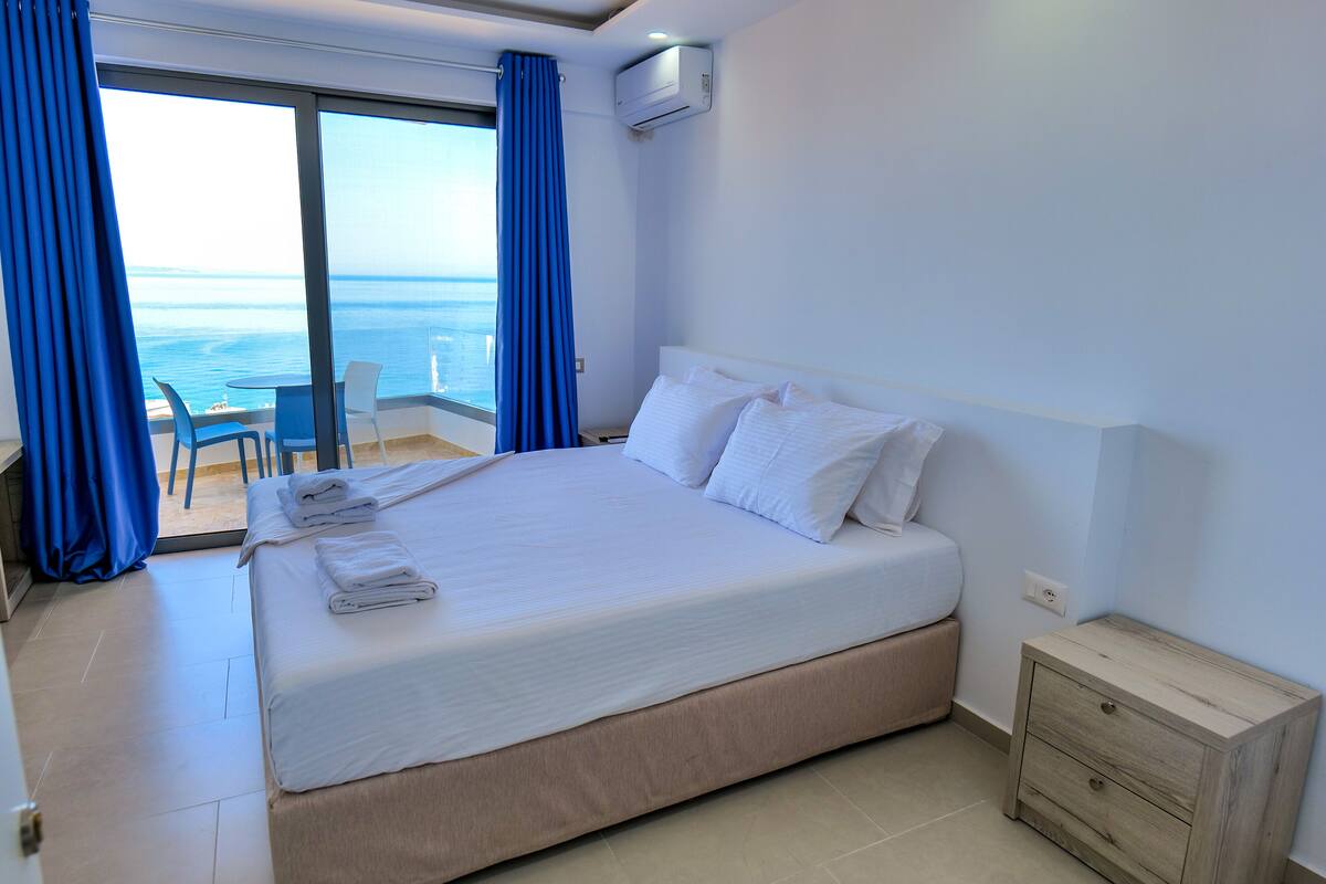 A hotel room with a large bed, a small bedside table, a view of the sea through a sliding glass door, and a balcony with chairs.