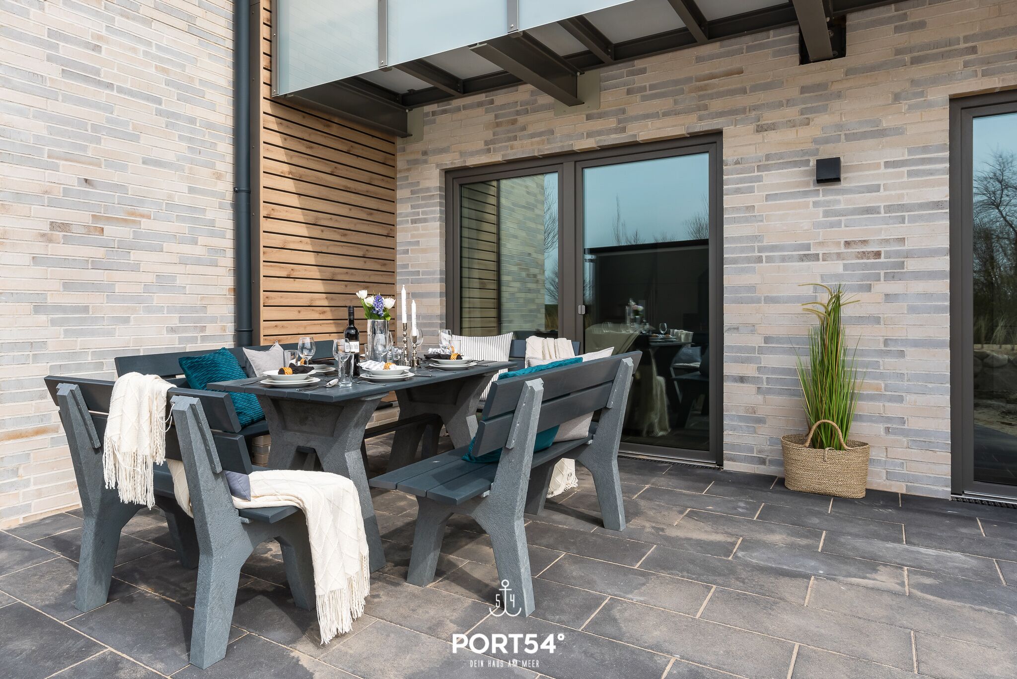 Une terrasse extérieure avec une table et des bancs, un pot de fleurs et une porte-fenêtre coulissante donnant sur un espace intérieur.