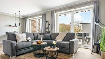 Un salon moderne avec un canapé d’angle, deux petites tables rondes et un coin repas visible à travers de grandes fenêtres.