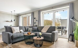 Een moderne woonkamer met een hoekbank, twee ronde salontafels en een eetgedeelte dat zichtbaar is door grote ramen.