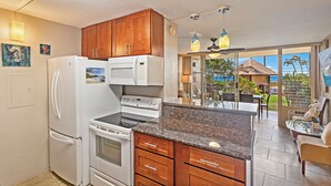 Une cuisine avec des meubles en bois, un réfrigérateur blanc, une cuisinière et un four à micro-ondes. On y trouve également un coin repas avec une table et des chaises, et une vue sur l’océan grâce à de grandes fenêtres.