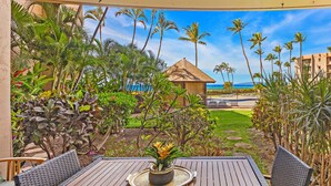 An outdoor patio with a wooden table and chairs, surrounded by tropical plants and a view of the ocean. - Breezy Maalaea Oceanfront Condo Newly Renovated Pool BBQ Near the Harbor (Wailuku)