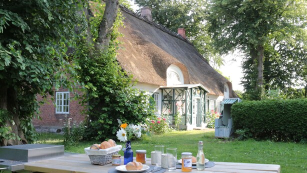 A picnic setup with a table, food, and drinks in a garden with a thatched-roof house in the background. - Apartment "PASTORATSGARTEN SCHLEI": Relax and enjoy (Norderbrarup)