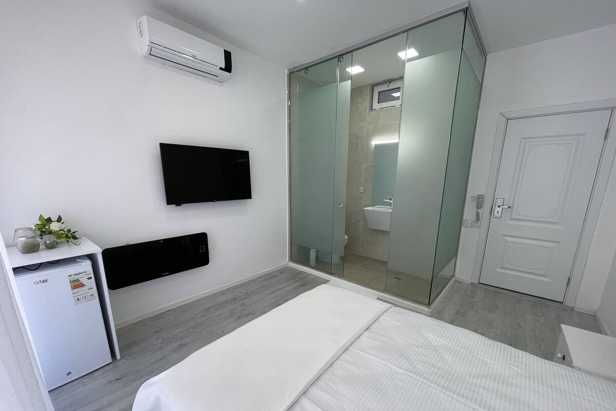 Une chambre d’hôtel moderne équipée d’un lit, d’un bureau avec un ordinateur, d’une télévision et d’une douche séparée par des parois en verre.