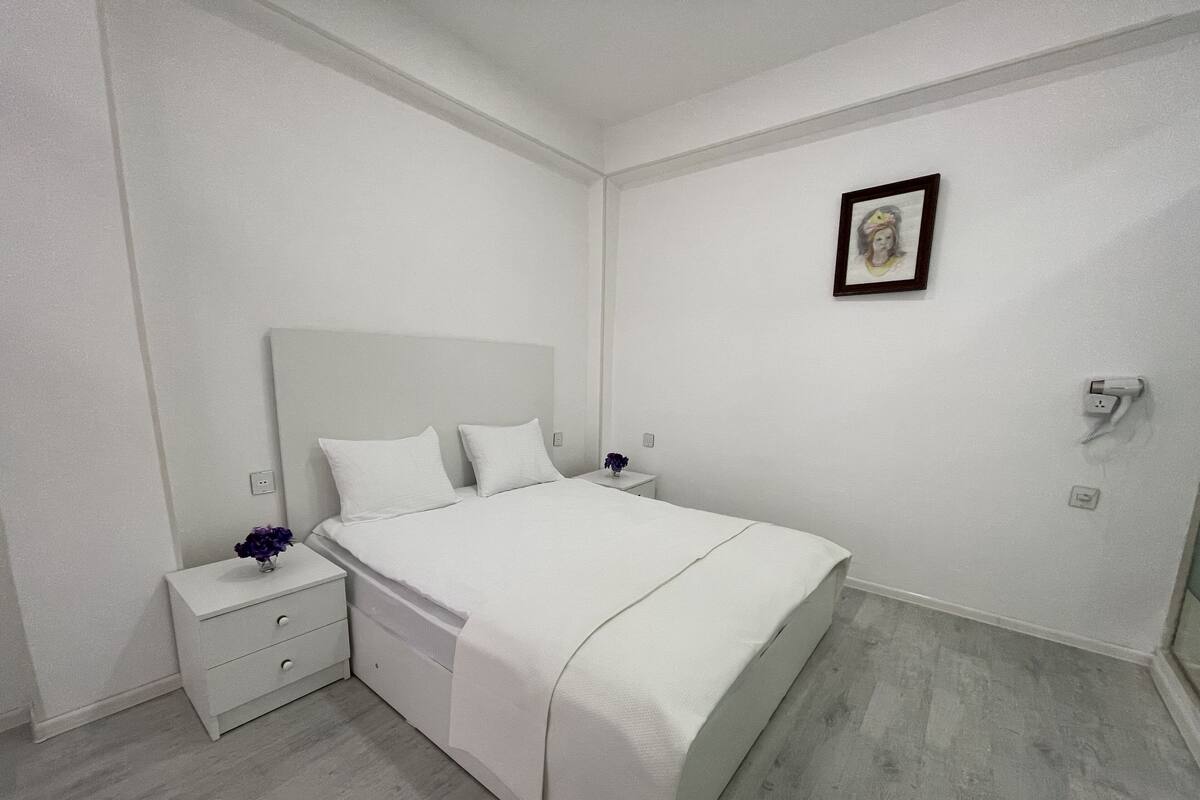 Une chambre à coucher avec un lit, des tables de chevet, une petite table sur laquelle est posé un vase, et un tableau encadré au mur.