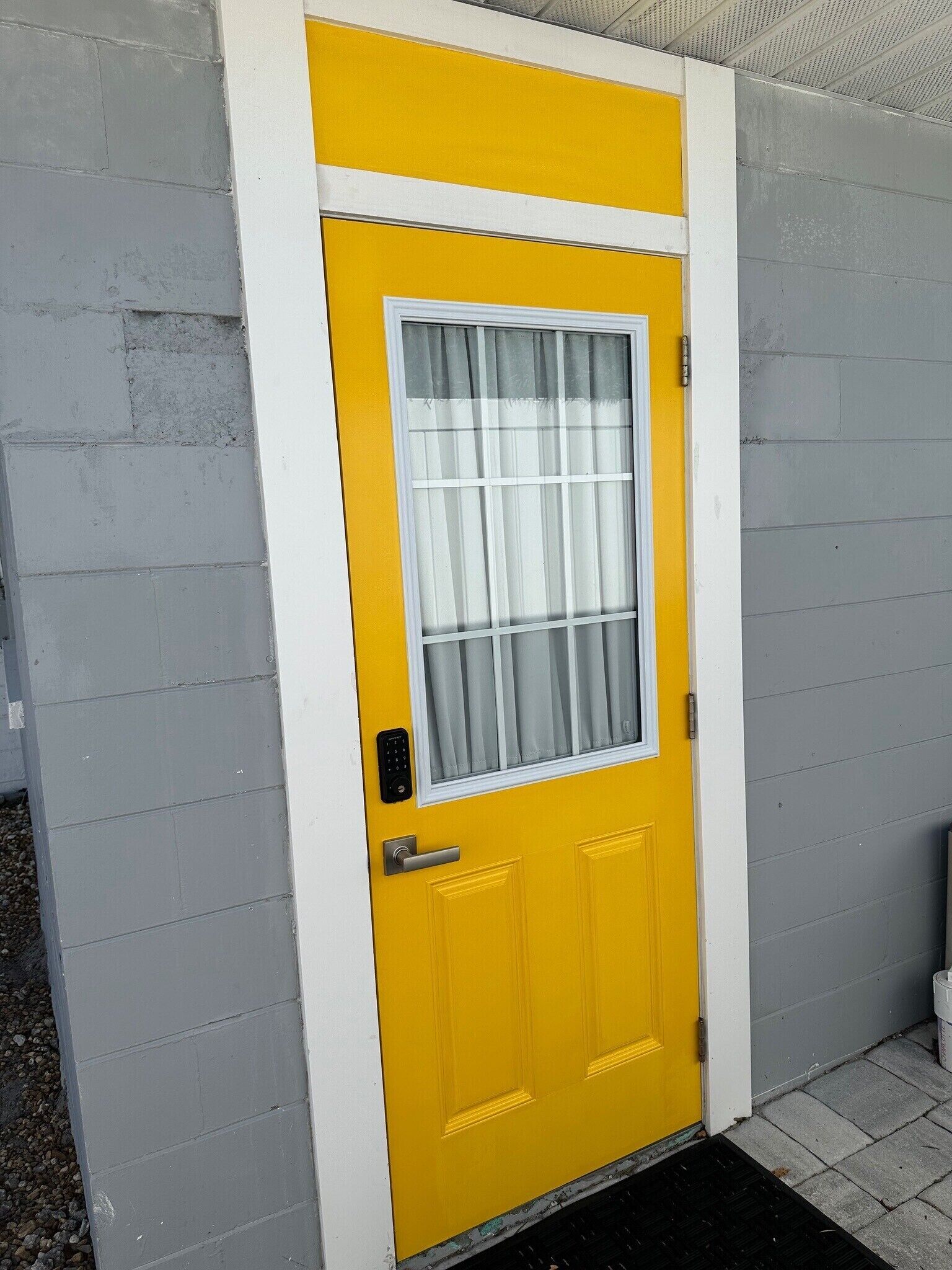 Una puerta amarilla con una ventana, un teclado electrónico para abrirla y un felpudo.