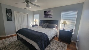 Ein Schlafzimmer mit Bett, Deckenventilator, Fenster mit Jalousien, Nachttisch und Wandbild.