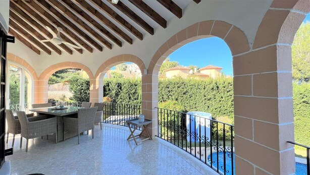 Un patio techado al aire libre con zona de comedor, que incluye una mesa y sillas, y vistas a una piscina y vegetación.
