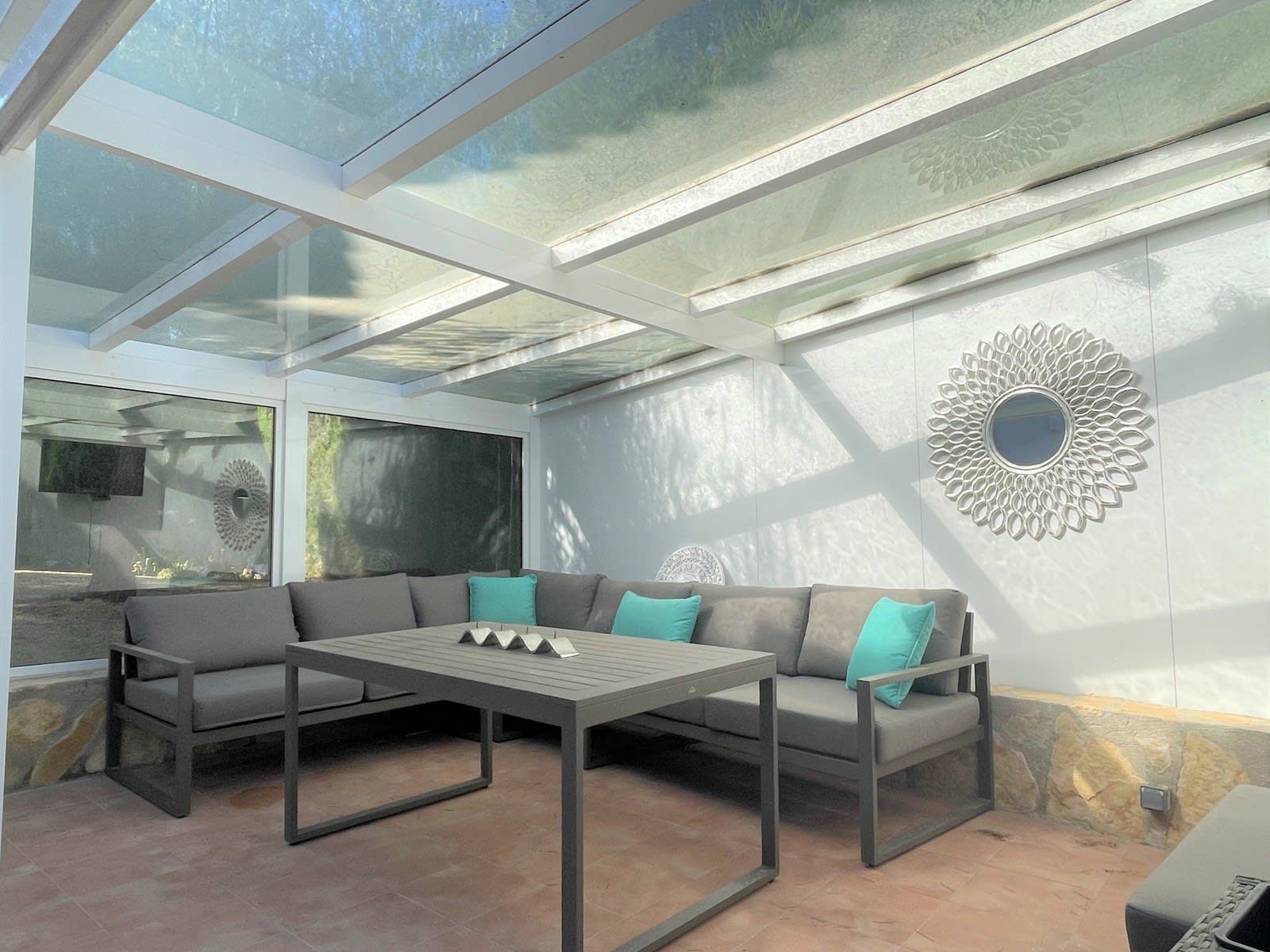 A sunroom with a glass ceiling, a sectional sofa with teal cushions, and a decorative wall mirror.