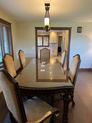 Une salle à manger avec une grande table en verre entourée de six chaises, un lustre et une vue sur la cuisine.