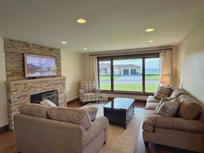 A living room with a stone fireplace, a television mounted above it, a large window with a view of a house and the ocean, and two sofas. - Lake View Ranch (Erie)