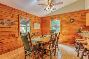 A wooden cabin interior with a dining table, chairs, a ceiling fan, and a window with a view of trees.