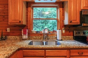 A kitchen with wooden cabinets, a granite countertop, a stainless steel sink, and a window with a forest view. - Miracle Mountain Cabin Secluded Mountain Getaway (Bryson City)