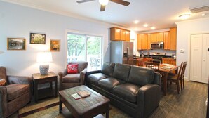 A living room with a leather sofa, a coffee table, and a dining area with a table and chairs. There is a kitchen with wooden cabinets and appliances.