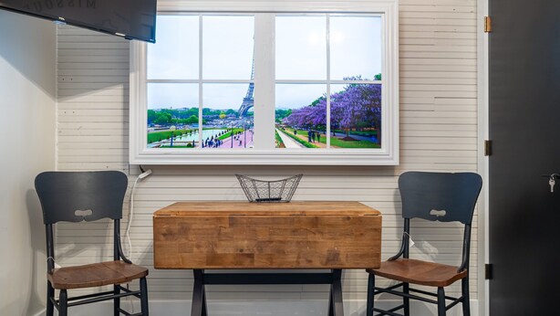 A dining area with a wooden table and two chairs, a window with a view of the Eiffel Tower, and a white wall with horizontal lines.