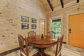 A wooden interior with a round table and chairs, framed pictures on the wall, and a window with a view of greenery.