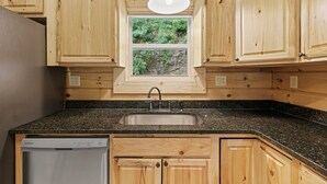 A kitchen with wooden cabinets, a granite countertop, a built-in dishwasher, and a window with a view of greenery.