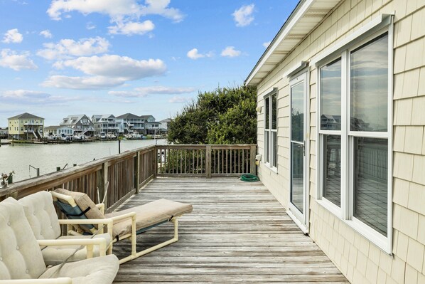 A wooden deck with a lounge chair, a view of the water, and a row of houses in the background.