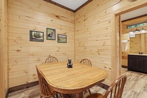 Une pièce aux murs lambrissés, avec une table et des chaises, des tableaux encadrés et un coin cuisine visible à travers une porte ouverte.