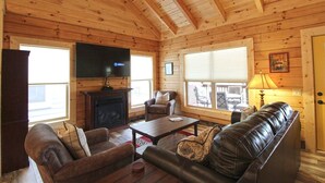 A cozy cabin interior with a fireplace, leather sofa, and a television mounted on the wall. - Moose Tracks Downtown Veranda Level Cabin with Hot Tub (Bryson City)