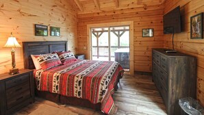 A wooden cabin bedroom with a bed, nightstands, a dresser, a television, and a window with a view. - Moose Tracks Downtown Veranda Level Cabin with Hot Tub (Bryson City)