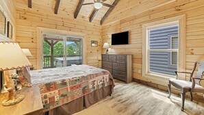 Un dormitorio de cabaña de madera con cama, cómoda, televisor y una ventana grande.