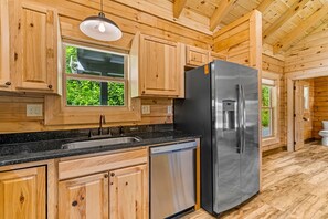 A kitchen with wooden cabinets, a stainless steel refrigerator, a dishwasher, and a window with a view of greenery.