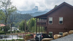 A two-story wooden house with a balcony, surrounded by a gravel driveway and a metal fence.
