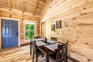 Un intérieur en bois avec une table à manger dressée pour quatre personnes, une porte bleue et une fenêtre donnant sur un paysage verdoyant.