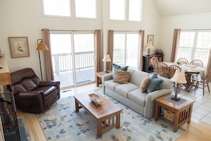 A spacious living area with a sofa, a coffee table, a brown leather chair, a dining table with chairs, and a balcony with curtains.