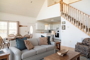 A living room with a sofa, a wooden coffee table, a patterned armchair, a dining table with chairs, and a kitchen in the background.