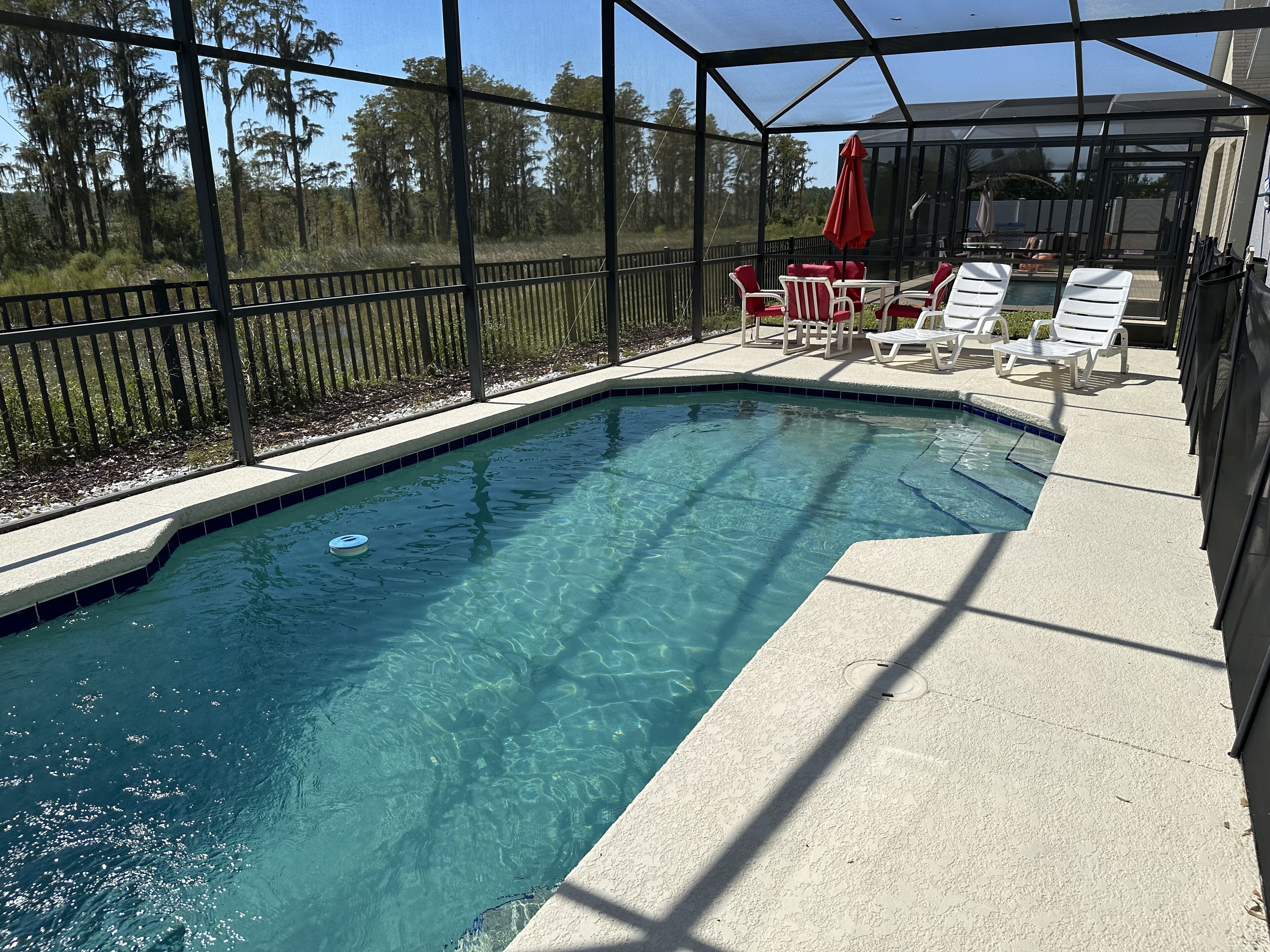 A swimming pool with clear water, surrounded by a covered patio area with lounge chairs and a red umbrella.
