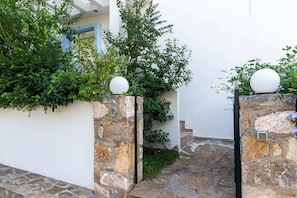 A stone pathway leading to a white building with a blue door, surrounded by greenery and stone walls.