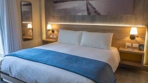 Hotellihuone, jossa on suuri sänky, kaksi yöpöytävalaisinta, yöpöytä ja seinälle ripustettu taulu.
