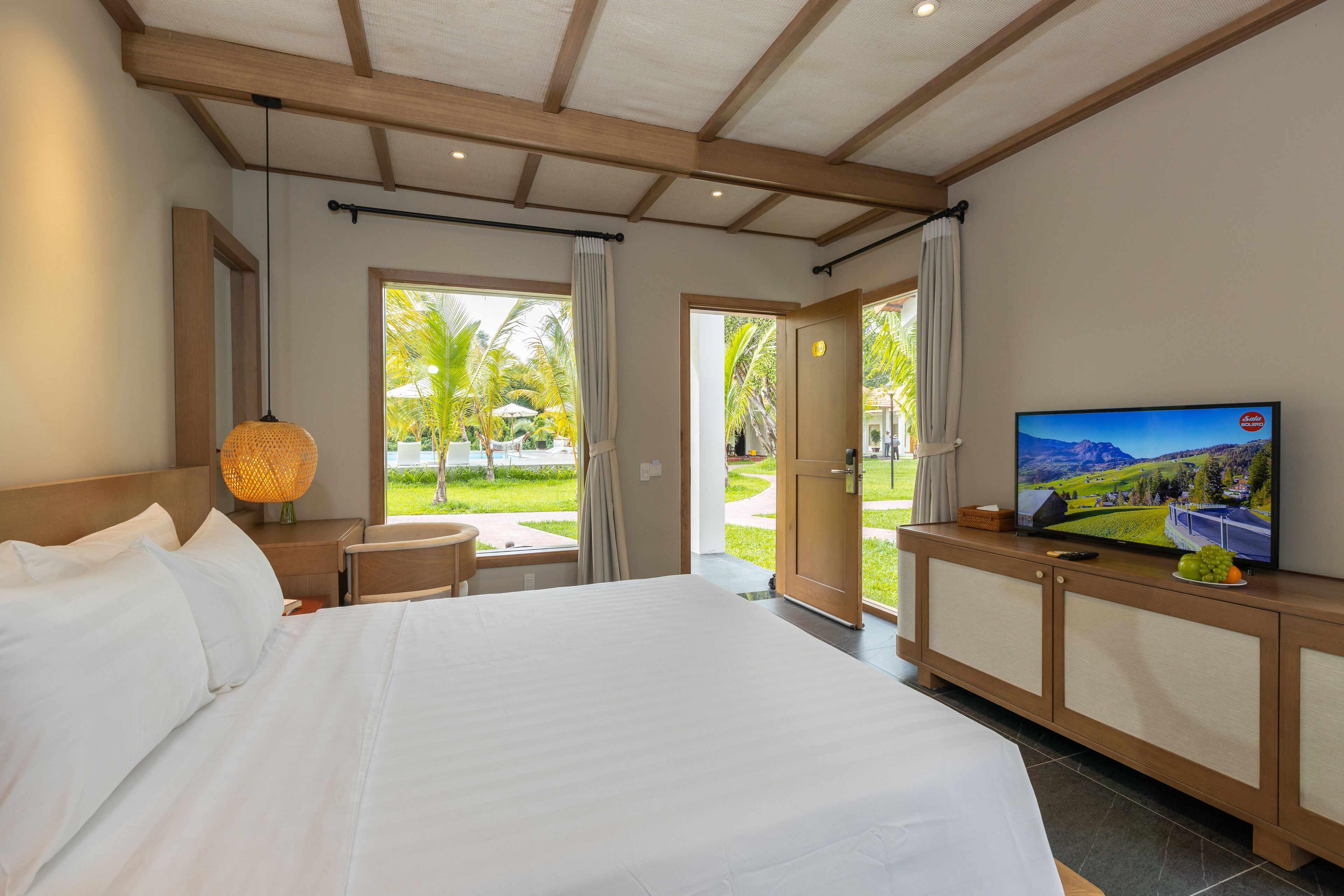 A hotel room with a large bed, a TV on a wooden console, a small table with a lamp, and a view of the outdoors through a glass door.
