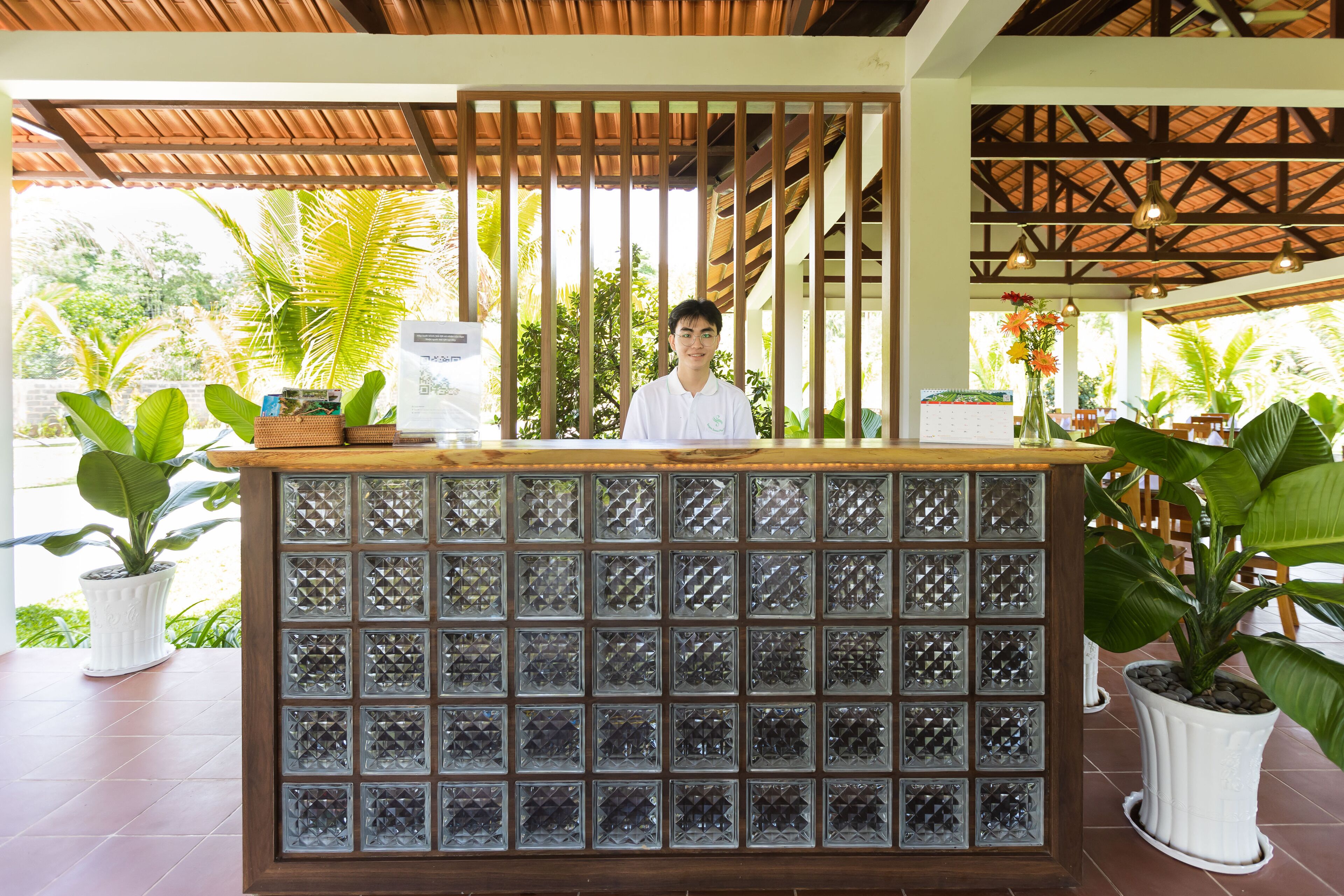 A reception desk with a wooden top and a glass panel design, surrounded by potted plants and a view of tropical vegetation.