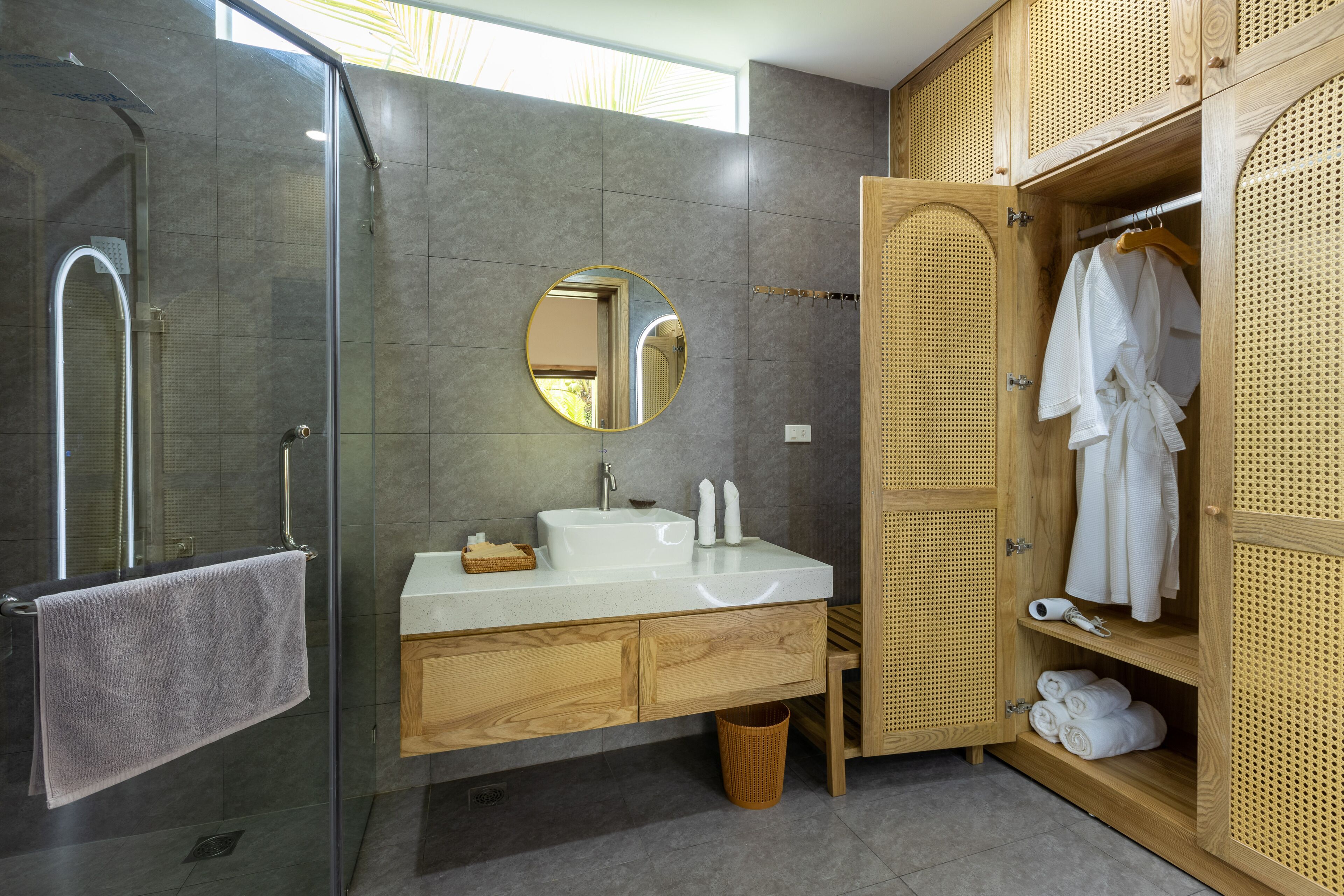 A modern bathroom with a glass-enclosed shower, a round mirror, a white sink, wooden cabinets, and a wooden wardrobe with a towel rack.