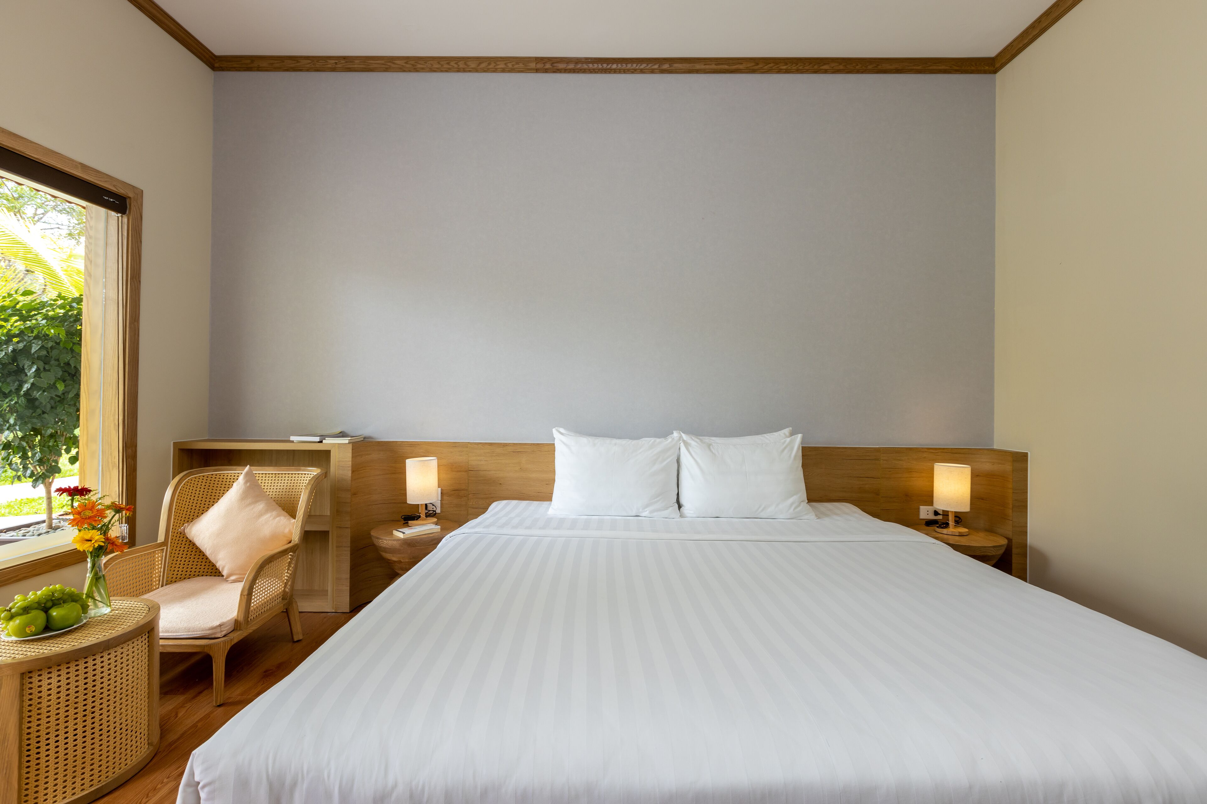Una habitación de hotel con una cama grande, dos mesitas de noche con lámparas, una silla de mimbre, una pequeña mesa con un florero y una ventana con vistas a zonas verdes.