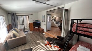 A compact living space with a sofa, a TV, a small table, a bunk bed, and a kitchenette in the background.