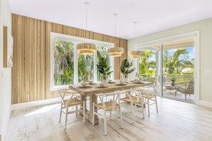 Uma área de refeições com mesa de madeira e cadeiras brancas, janelas amplas com vista para palmeiras e luminárias pendentes.
