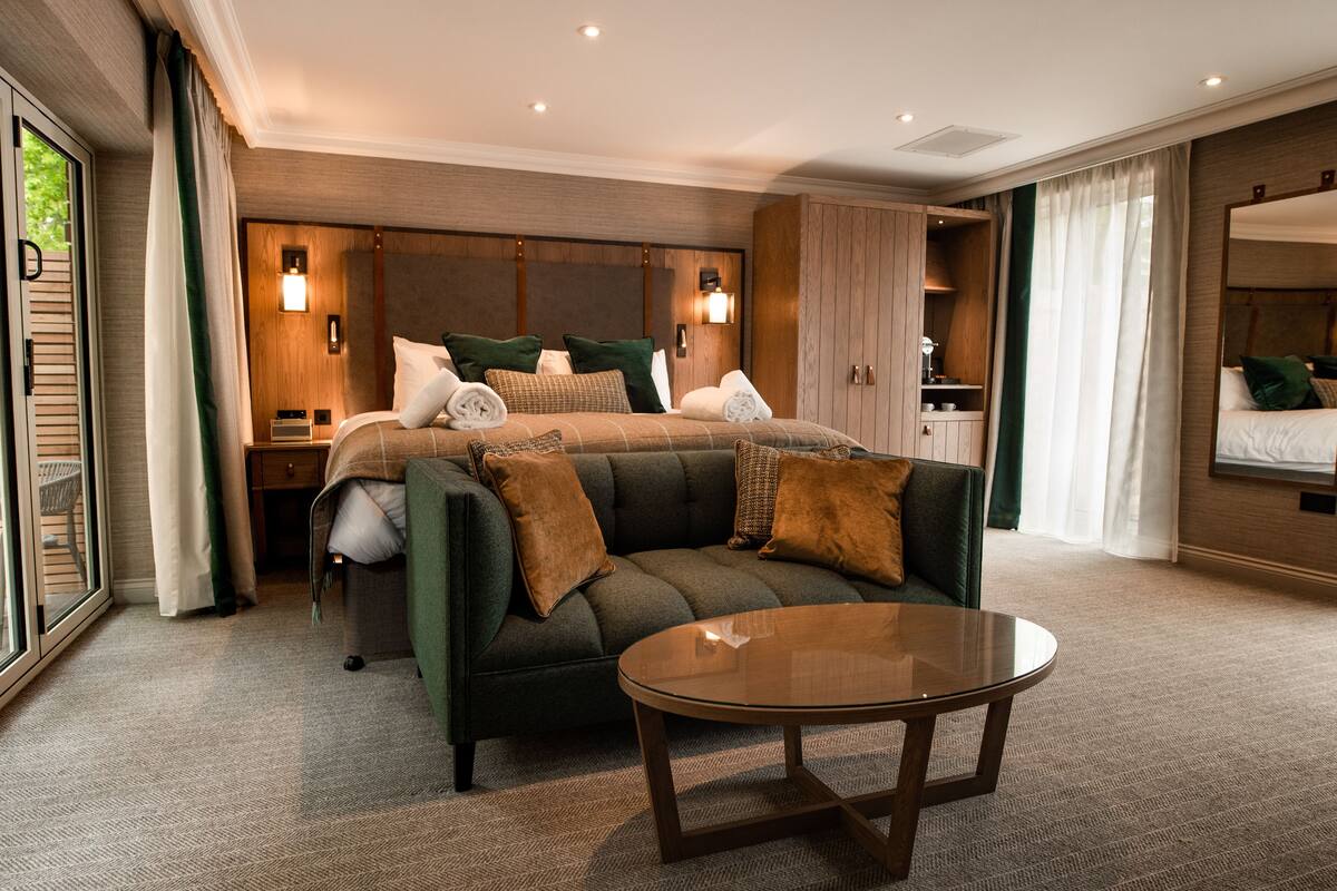 A modern hotel room with a large bed, a sofa, a glass coffee table, and a view of greenery through the window.