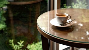 Eine Tasse Kaffee auf einem Glastisch mit einem Löffel, nahe einem Fenster mit Blick auf einen Wald.